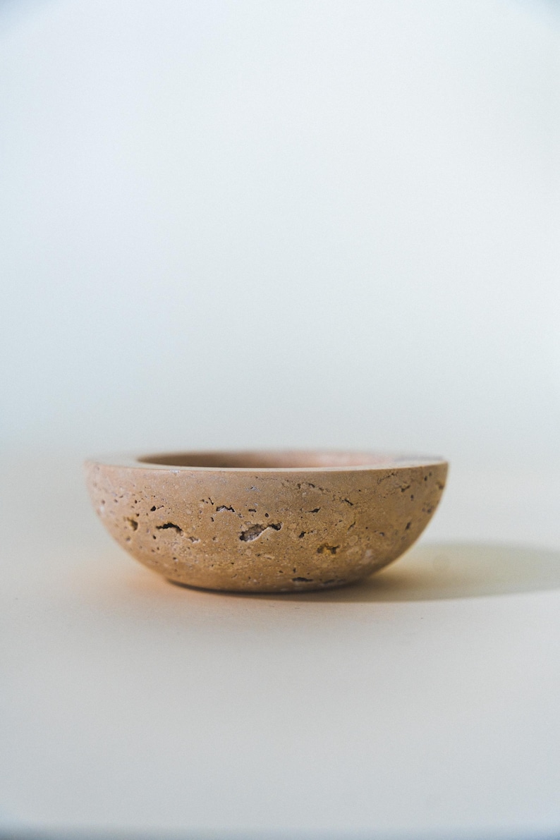 May include: A small, round, beige bowl with a textured surface. The bowl has a shallow depth and a wide rim. The neutral color and simple design make it suitable for various uses, such as a decorative piece or a small serving dish.