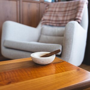 May include: A small, round, off-white ashtray with a cigar resting inside, placed on a wooden table. The table has a glossy finish and a warm, reddish-brown color. A light-colored armchair and a wooden cabinet are in the background.