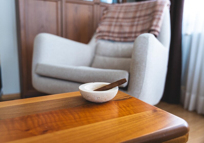 May include: A small, round, off-white ashtray with a cigar resting inside, placed on a wooden table. The table has a glossy finish and a warm, reddish-brown color. A light-colored armchair and a wooden cabinet are in the background.