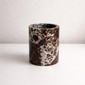 May include: A cylindrical container made of marble. The marble has a pattern of dark brown and white spots and veins. The container is a solid, sturdy design, suitable for holding items.