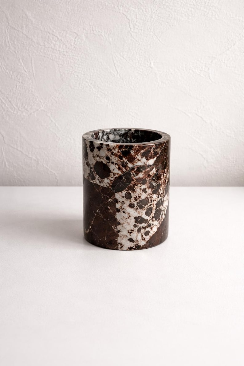 May include: A cylindrical container made of marble. The marble has a pattern of dark brown and white spots and veins. The container is a solid, sturdy design, suitable for holding items.