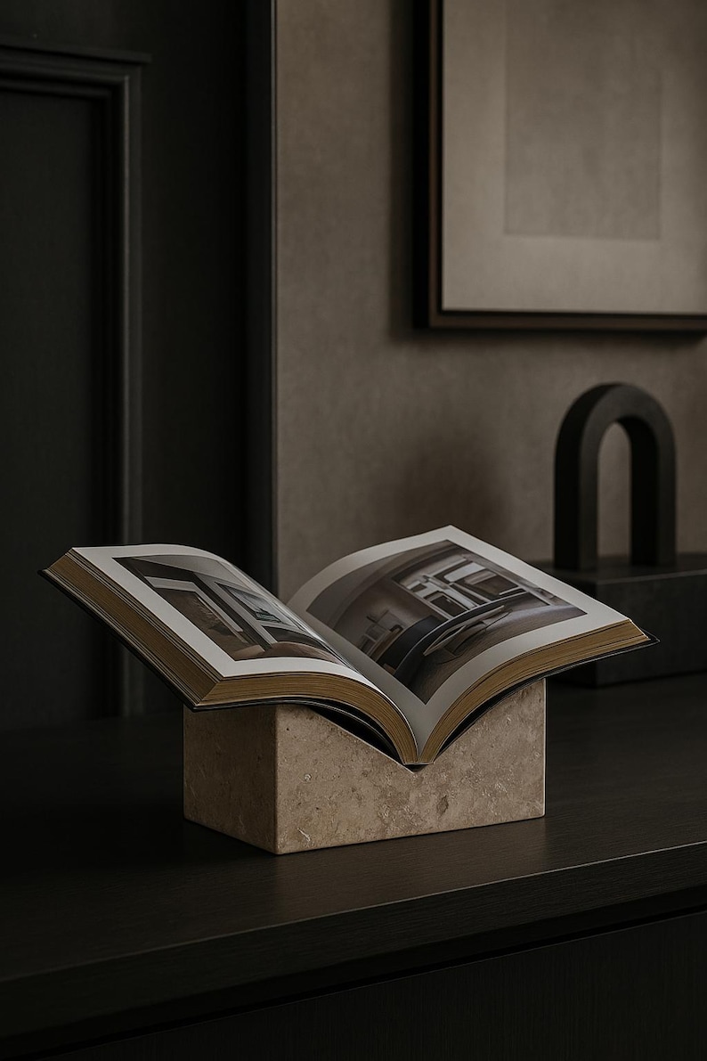 May include: An open book with gold-edged pages rests on a square, beige stone base. The book displays interior photographs. The scene is set against a dark, neutral background with a framed picture and a dark sculpture.