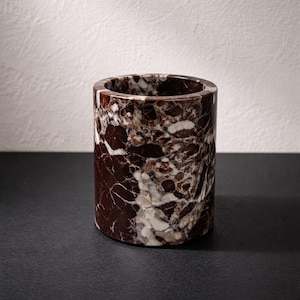 May include: A cylindrical container made of marble. The marble has a dark brown base color with white and light brown veining throughout. The container is sitting on a black surface.