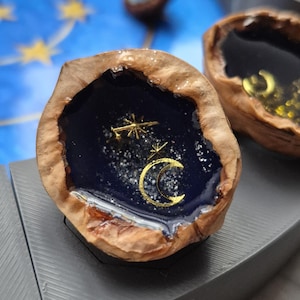 Könnte beinhalten: Eine Walnussschale mit einem dunkelblauen Harzinnenraum, verziert mit goldfarbenen Sternen und einem Mond. Die Schale befindet sich auf einer grauen Oberfläche, mit einer weiteren ähnlichen Schale im Hintergrund. Das Harz hat einen Glitzereffekt.