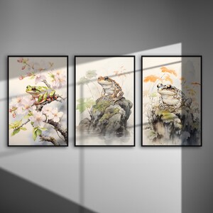 May include: Set of three framed watercolor prints featuring frogs. One print shows a green frog on a flowering branch, another a frog on a rock, and the third a frog with autumn leaves. The art has a soft, natural aesthetic.