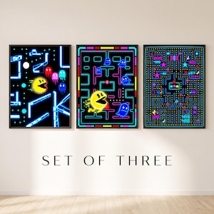Peut inclure: Un ensemble de trois affiches murales encadrées présentant des graphismes de jeux vidéo pixélisés. Les impressions montrent un personnage circulaire jaune naviguant dans des labyrinthes bleu néon et rose avec des fantômes, des fruits et des scores.