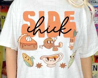 Può includere: Maglietta bianca con il testo "Side chick" in lettere arancioni. La maglietta presenta un'illustrazione di cartoni animati di una torta, una pannocchia di mais, una carota, una salsiera e un fagiolino.