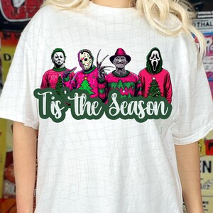 May include: White t-shirt with a graphic of four horror movie characters wearing Christmas sweaters. The text "Tis' the Season" is printed below the characters.