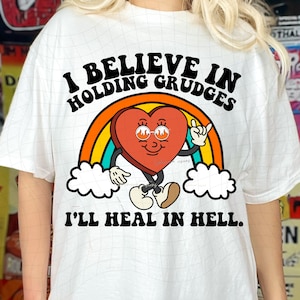 Puede incluir: Camiseta blanca con un gráfico de un corazón rojo con gafas de sol y un arcoíris. El texto "I believe in holding grudges I'll heal in hell." está impreso en la camiseta.