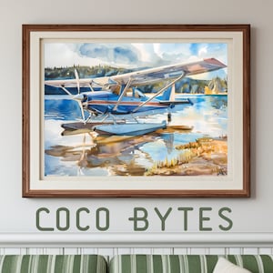 May include: Watercolor painting of a blue and white seaplane on a lake, framed in a brown wooden frame. The artwork reflects the plane and the surrounding landscape in the water. The text "COCO BYTES" is at the bottom of the frame.