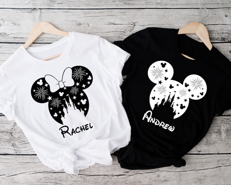 Puede incluir: Dos camisetas blancas con orejas de Mickey Mouse negras y un dise&ntilde;o de castillo. Una camiseta dice "Rachel" y la otra dice "Andrew".