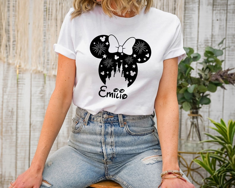 Puede incluir: Camiseta blanca con un gr&aacute;fico en blanco y negro de orejas de Mickey Mouse con un lazo, fuegos artificiales y un castillo en el interior. El texto "Emilio" est&aacute; impreso debajo del gr&aacute;fico.