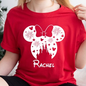 Puede incluir: Camiseta roja con una silueta blanca de orejas de Mickey Mouse con un lazo y fuegos artificiales dentro de las orejas. La silueta tambi&eacute;n incluye un castillo y el texto "Rachel".