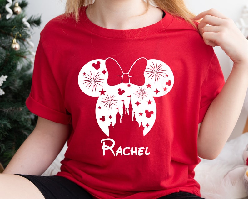 Puede incluir: Camiseta roja con una silueta blanca de orejas de Mickey Mouse con un lazo y fuegos artificiales dentro de las orejas. La silueta tambi&eacute;n incluye un castillo y el texto "Rachel".