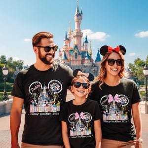 K&ouml;nnte beinhalten: Drei Personen tragen schwarze T-Shirts mit einem Disneyland-Design vor einem Schloss. Die Shirts tragen die Aufschrift "Daddy", "Mommy" und "Nora" &uuml;ber dem Disneyland-Logo. Die Shirts tragen auch die Aufschrift "Family Vacation 2026".