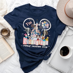K&ouml;nnte beinhalten: Marineblaues T-Shirt mit Disneyland-Design, das ein Schloss, Feuerwerk und Disney-Figuren zeigt. Das Shirt enth&auml;lt den Namen "Tim" und den Text "Family Vacation 2026". Ein Hut und eine Kaffeetasse sind im Hintergrund zu sehen.