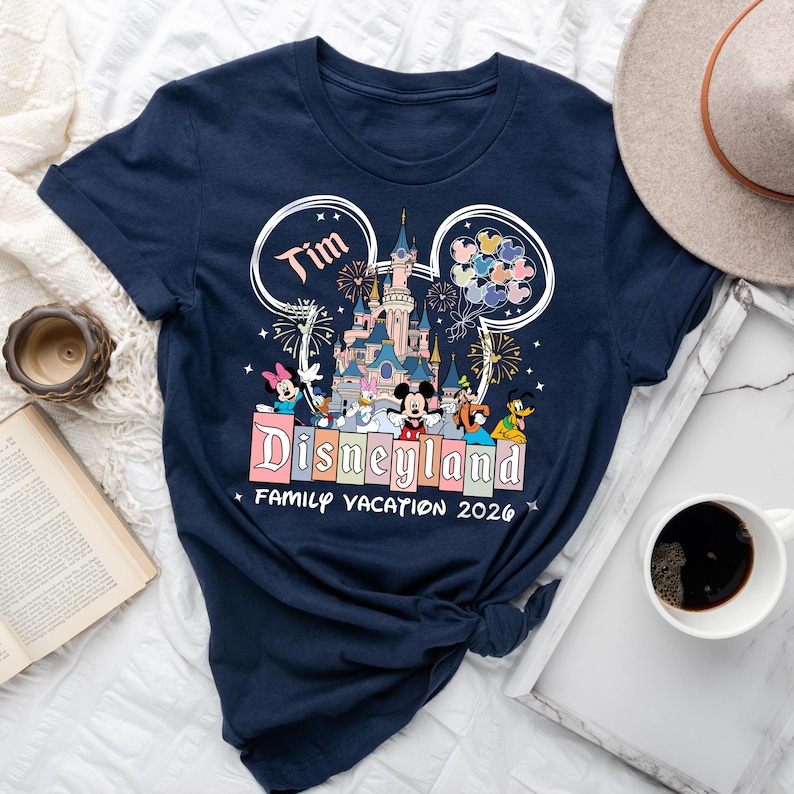 K&ouml;nnte beinhalten: Marineblaues T-Shirt mit Disneyland-Design, das ein Schloss, Feuerwerk und Disney-Figuren zeigt. Das Shirt enth&auml;lt den Namen "Tim" und den Text "Family Vacation 2026". Ein Hut und eine Kaffeetasse sind im Hintergrund zu sehen.