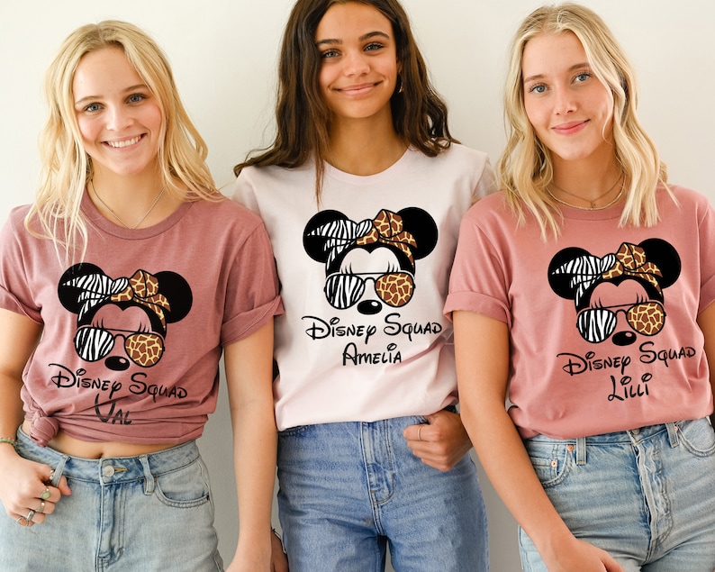 May include: Three pink and white t-shirts with a cartoon image of Mickey Mouse wearing sunglasses and a zebra and cheetah print headband. The text on the shirts reads "Disney Squad" with a name below.
