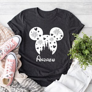 Puede incluir: Una camiseta gris oscuro con una silueta blanca de la cabeza de Mickey Mouse. La silueta incluye un castillo, fuegos artificiales y estrellas. El nombre "ANDREW" est&aacute; escrito debajo de la silueta.