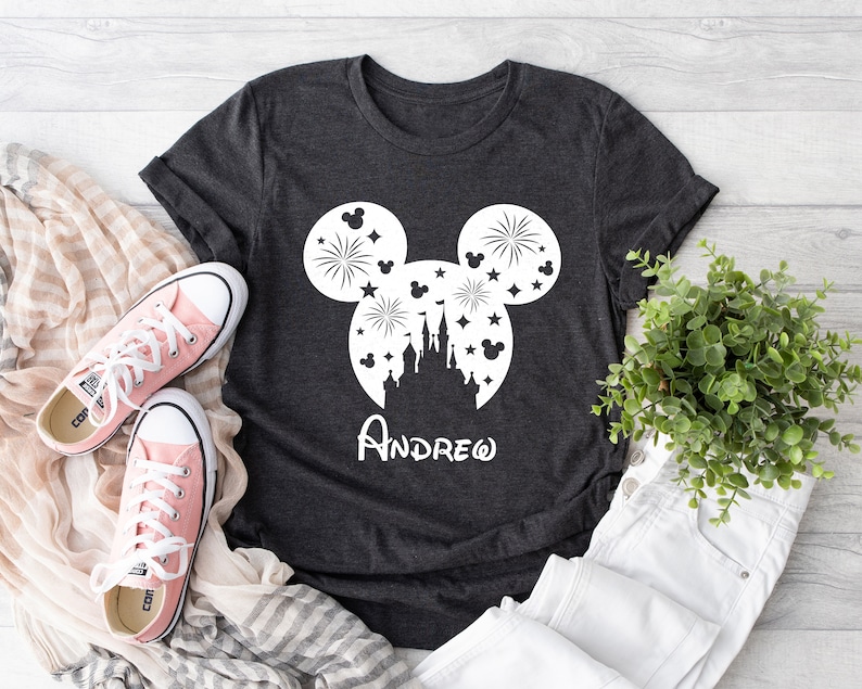 Puede incluir: Una camiseta gris oscuro con una silueta blanca de la cabeza de Mickey Mouse. La silueta incluye un castillo, fuegos artificiales y estrellas. El nombre "ANDREW" est&aacute; escrito debajo de la silueta.