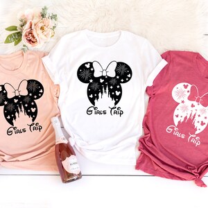 Puede incluir: Tres camisetas con una silueta en blanco y negro de orejas de Mickey Mouse con un lazo y un castillo dentro de las orejas. El texto "Girls Trip" est&aacute; debajo de las orejas. Las camisetas son de color melocot&oacute;n, blanco y rojo.