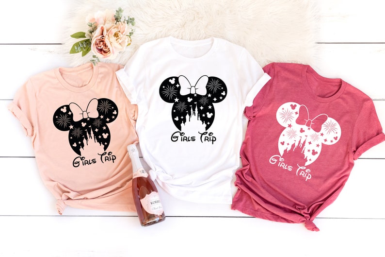 Puede incluir: Tres camisetas con una silueta en blanco y negro de orejas de Mickey Mouse con un lazo y un castillo dentro de las orejas. El texto "Girls Trip" est&aacute; debajo de las orejas. Las camisetas son de color melocot&oacute;n, blanco y rojo.