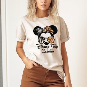 May include: A white t-shirt with a black and white zebra print Minnie Mouse head wearing sunglasses with a brown and orange giraffe print. The text "Disney Trip Cousin" is printed below the Minnie Mouse head.