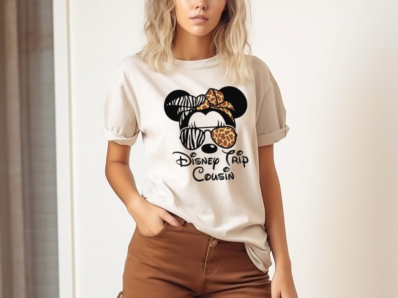May include: A white t-shirt with a black and white zebra print Minnie Mouse head wearing sunglasses with a brown and orange giraffe print. The text "Disney Trip Cousin" is printed below the Minnie Mouse head.