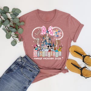 K&ouml;nnte beinhalten: Staubrosa T-Shirt mit Disneyland-Grafik, die ein Schloss, Charaktere und den Text "Aunt Disneyland Family Vacation 2026" zeigt. Das Design beinhaltet eine rosa Schleife und Ballons. Blaue Jeans und Sandalen sind ebenfalls zu sehen.