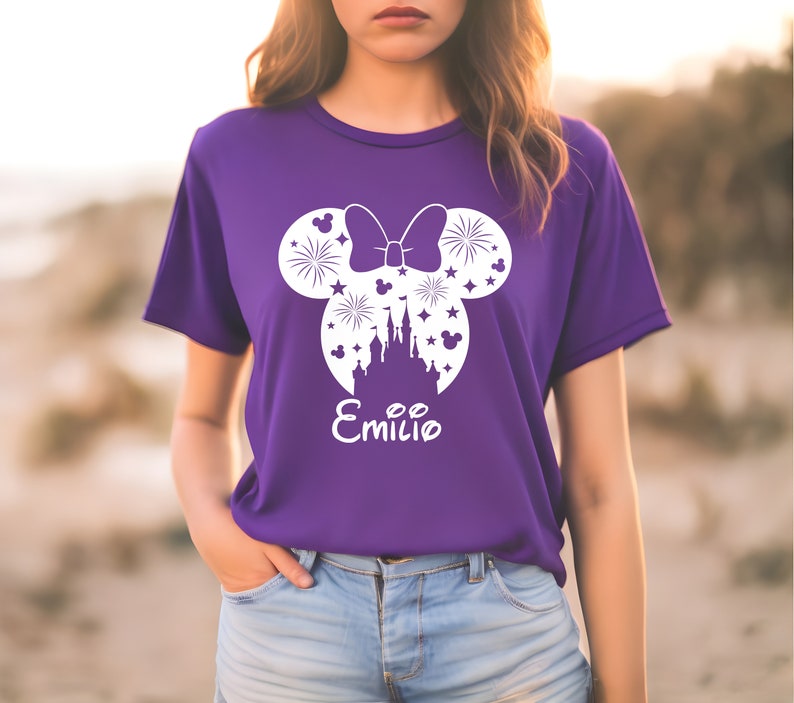 Puede incluir: Camiseta morada con una silueta blanca de orejas de Mickey Mouse, un lazo, fuegos artificiales, un castillo y estrellas. El texto "Emilio" est&aacute; debajo del dise&ntilde;o.