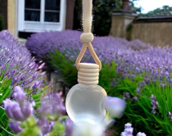 Può includere: Un profumatore per auto in vetro bianco a forma di mela con un tappo in legno e una corda bianca che pende. Il profumatore è appeso davanti a un campo di fiori di lavanda viola.