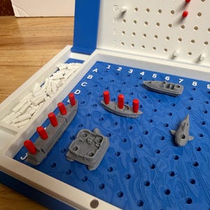 Peut inclure: Un jeu de soci&eacute;t&eacute; Battleship dans un &eacute;tui en plastique bleu et blanc. Le plateau de jeu pr&eacute;sente un motif d'oc&eacute;an bleu avec des navires gris et des chevilles rouges. Le jeu comprend divers mod&egrave;les de navires et des chevilles pour marquer les coups et les manques.