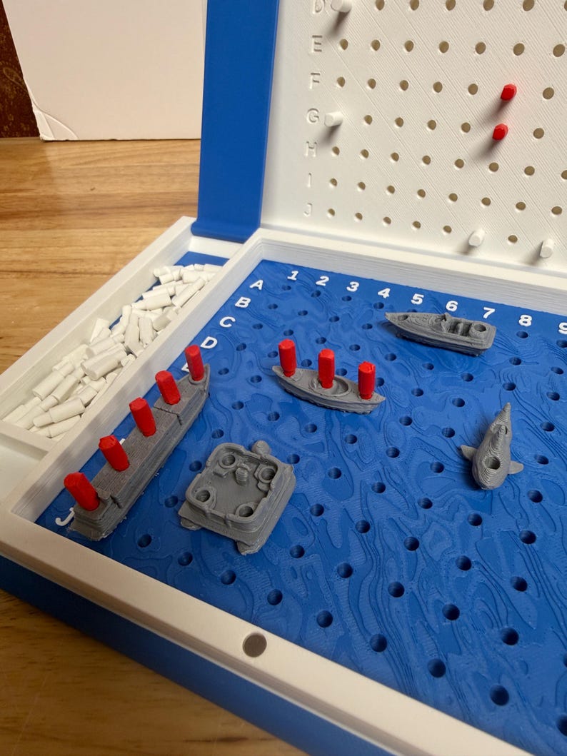 Peut inclure: Un jeu de soci&eacute;t&eacute; Battleship dans un &eacute;tui en plastique bleu et blanc. Le plateau de jeu pr&eacute;sente un motif d'oc&eacute;an bleu avec des navires gris et des chevilles rouges. Le jeu comprend divers mod&egrave;les de navires et des chevilles pour marquer les coups et les manques.