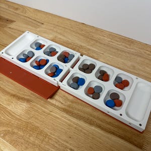 Peut inclure: Un jeu de soci&eacute;t&eacute; Mancala blanc et orange avec des pierres color&eacute;es. Le plateau de jeu comporte deux rang&eacute;es de six poches et deux grandes poches d'extr&eacute;mit&eacute;. Les pierres sont rouges, bleues, marron et grises.