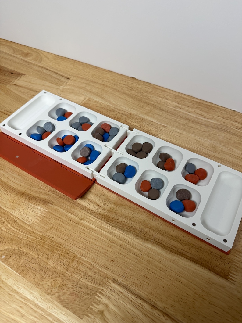 Peut inclure: Un jeu de soci&eacute;t&eacute; Mancala blanc et orange avec des pierres color&eacute;es. Le plateau de jeu comporte deux rang&eacute;es de six poches et deux grandes poches d'extr&eacute;mit&eacute;. Les pierres sont rouges, bleues, marron et grises.