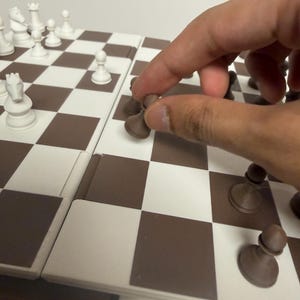 Peut inclure: Un jeu d'&eacute;checs avec un &eacute;chiquier &agrave; carreaux marron et blanc. Des pi&egrave;ces d'&eacute;checs blanches sont dispos&eacute;es sur le plateau, avec une main d&eacute;pla&ccedil;ant un pion marron. Les pi&egrave;ces d'&eacute;checs comprennent un roi, une reine, une tour, un cavalier, un fou et des pions.