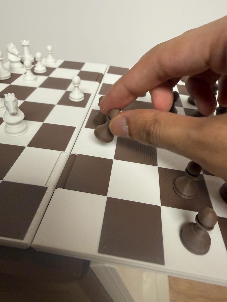 Peut inclure: Un jeu d'&eacute;checs avec un &eacute;chiquier &agrave; carreaux marron et blanc. Des pi&egrave;ces d'&eacute;checs blanches sont dispos&eacute;es sur le plateau, avec une main d&eacute;pla&ccedil;ant un pion marron. Les pi&egrave;ces d'&eacute;checs comprennent un roi, une reine, une tour, un cavalier, un fou et des pions.