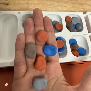 Peut inclure: Une main tenant plusieurs pi&egrave;ces de jeu color&eacute;es, lisses et en forme de pierre. Les pi&egrave;ces sont orange, bleu, gris et marron, avec une surface textur&eacute;e. Un plateau de jeu blanc et orange avec des compartiments est visible en arri&egrave;re-plan.