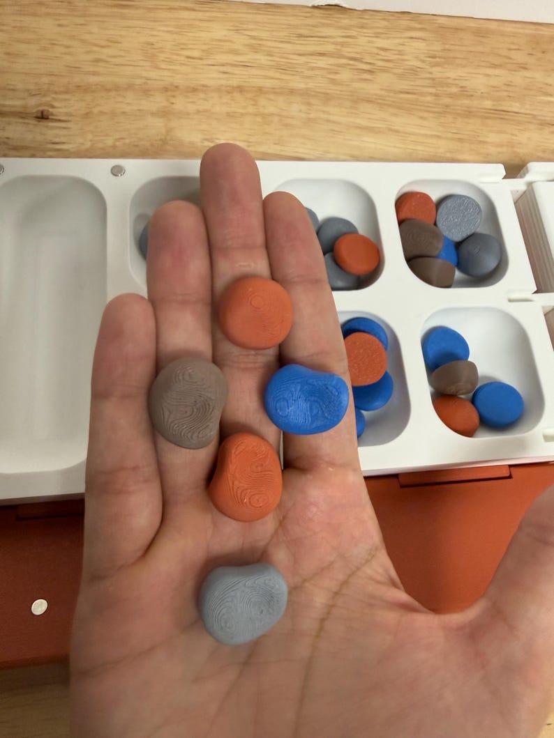 Peut inclure: Une main tenant plusieurs pi&egrave;ces de jeu color&eacute;es, lisses et en forme de pierre. Les pi&egrave;ces sont orange, bleu, gris et marron, avec une surface textur&eacute;e. Un plateau de jeu blanc et orange avec des compartiments est visible en arri&egrave;re-plan.
