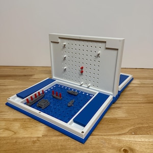 Peut inclure: Un jeu de soci&eacute;t&eacute; Battleship bleu et blanc avec des mod&egrave;les de navires miniatures. Le plateau de jeu ouvert r&eacute;v&egrave;le une surface de jeu bleue avec une grille et des mod&egrave;les de navires. Le couvercle blanc comporte des trous pour les chevilles et des chevilles rouges ins&eacute;r&eacute;es.