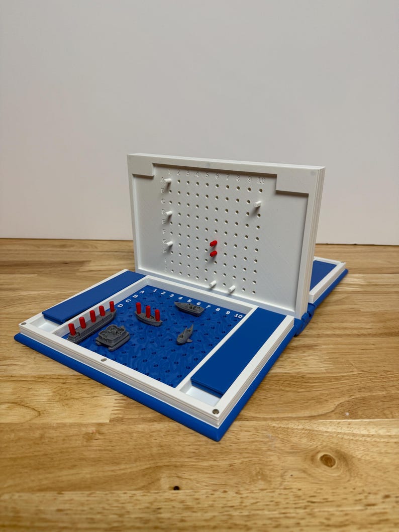 Peut inclure: Un jeu de soci&eacute;t&eacute; Battleship bleu et blanc avec des mod&egrave;les de navires miniatures. Le plateau de jeu ouvert r&eacute;v&egrave;le une surface de jeu bleue avec une grille et des mod&egrave;les de navires. Le couvercle blanc comporte des trous pour les chevilles et des chevilles rouges ins&eacute;r&eacute;es.