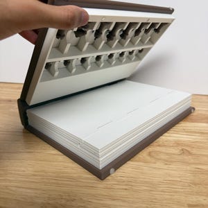 Peut inclure: Un organisateur en forme de livre marron et blanc, ouvert pour r&eacute;v&eacute;ler de petits compartiments. La partie sup&eacute;rieure comporte plusieurs fentes et la partie inf&eacute;rieure contient une pile de pages blanches. L'organisateur est sur une surface en bois.