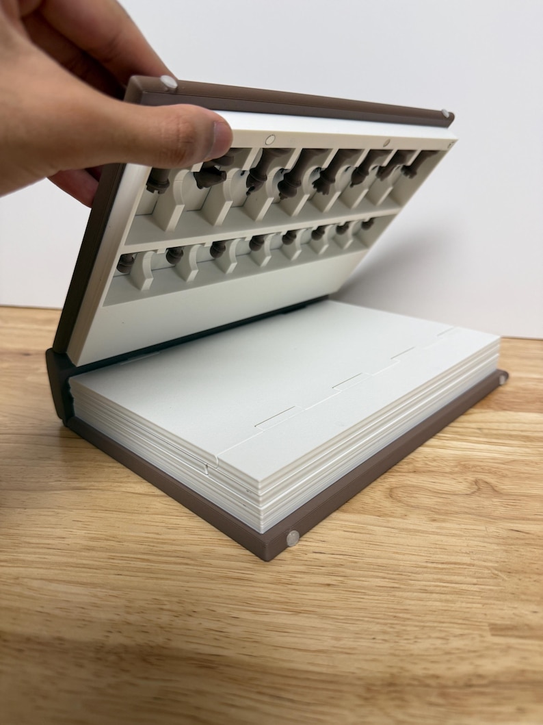 Peut inclure: Un organisateur en forme de livre marron et blanc, ouvert pour r&eacute;v&eacute;ler de petits compartiments. La partie sup&eacute;rieure comporte plusieurs fentes et la partie inf&eacute;rieure contient une pile de pages blanches. L'organisateur est sur une surface en bois.