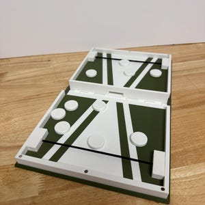 Peut inclure: Un jeu de soci&eacute;t&eacute; blanc et vert avec un design &agrave; charni&egrave;re. Le jeu pr&eacute;sente une surface de jeu verte avec des lignes blanches et des pi&egrave;ces de jeu circulaires. Le jeu est ouvert, r&eacute;v&eacute;lant l'aire de jeu et les pi&egrave;ces, pos&eacute; sur une surface en bois.