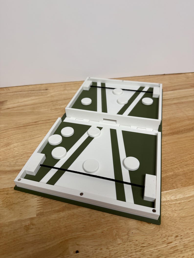 Peut inclure: Un jeu de soci&eacute;t&eacute; blanc et vert avec un design &agrave; charni&egrave;re. Le jeu pr&eacute;sente une surface de jeu verte avec des lignes blanches et des pi&egrave;ces de jeu circulaires. Le jeu est ouvert, r&eacute;v&eacute;lant l'aire de jeu et les pi&egrave;ces, pos&eacute; sur une surface en bois.