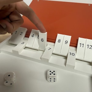 Peut inclure: Un jeu de soci&eacute;t&eacute; blanc avec des tuiles num&eacute;rot&eacute;es et des d&eacute;s. Le jeu a un dessus rouge et une base blanche. Un doigt pointe vers la tuile num&eacute;ro 6. Deux d&eacute;s sont sur la surface blanche.