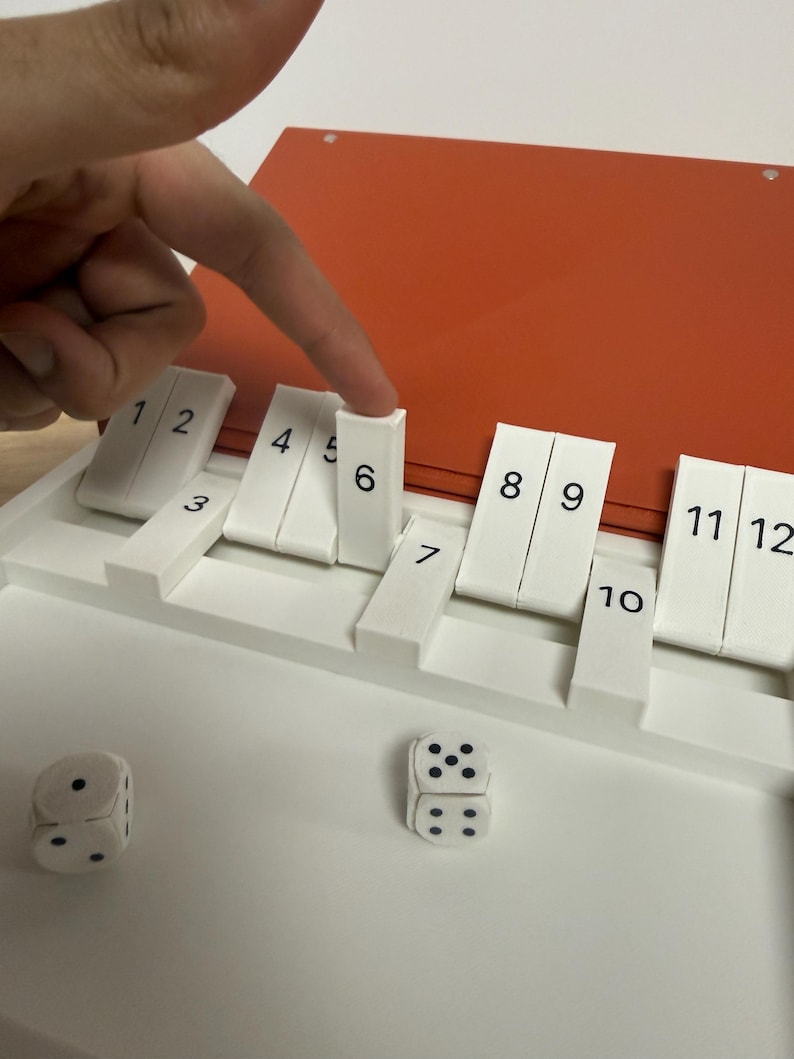 Peut inclure: Un jeu de soci&eacute;t&eacute; blanc avec des tuiles num&eacute;rot&eacute;es et des d&eacute;s. Le jeu a un dessus rouge et une base blanche. Un doigt pointe vers la tuile num&eacute;ro 6. Deux d&eacute;s sont sur la surface blanche.