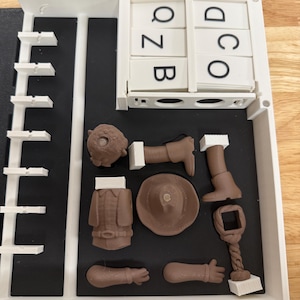 Peut inclure: Un organisateur en plastique blanc avec des compartiments, contenant des pi&egrave;ces marron imprim&eacute;es en 3D d'une figurine, dont un chapeau, un manteau et des membres. L'organisateur contient &eacute;galement des tuiles de lettres avec les lettres Q, Z, B, D, O et C.