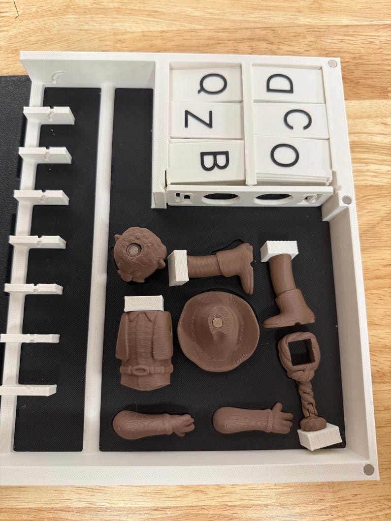 Peut inclure: Un organisateur en plastique blanc avec des compartiments, contenant des pi&egrave;ces marron imprim&eacute;es en 3D d'une figurine, dont un chapeau, un manteau et des membres. L'organisateur contient &eacute;galement des tuiles de lettres avec les lettres Q, Z, B, D, O et C.