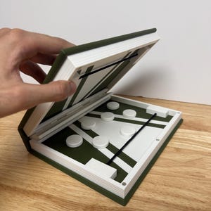 Peut inclure: Un jeu de soci&eacute;t&eacute; vert et blanc dans un &eacute;tui en forme de livre. Le plateau de jeu pr&eacute;sente un motif vert et blanc avec des lignes noires et des pions blancs. L'&eacute;tui est ouvert, r&eacute;v&eacute;lant le jeu.